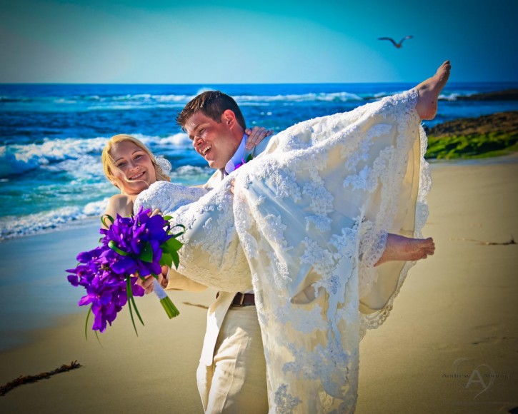 Groom carrying bride with purple wedding bouquet blue ocean and birds flying by San Diego Wedding Photographer Andrew Abouna